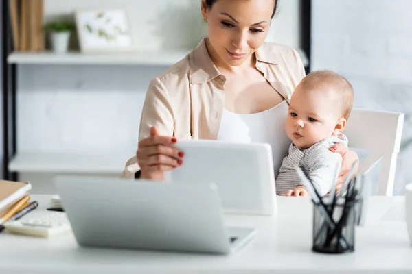 Enfoque selectivo de la madre sosteniendo la tableta digital cerca del niño y el ordenador portátil - foto de stock