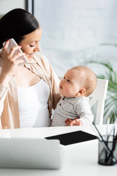 Enfoque selectivo de la madre feliz sosteniendo en brazos hijo lactante y hablando en el teléfono inteligente cerca de gadgets - foto de stock