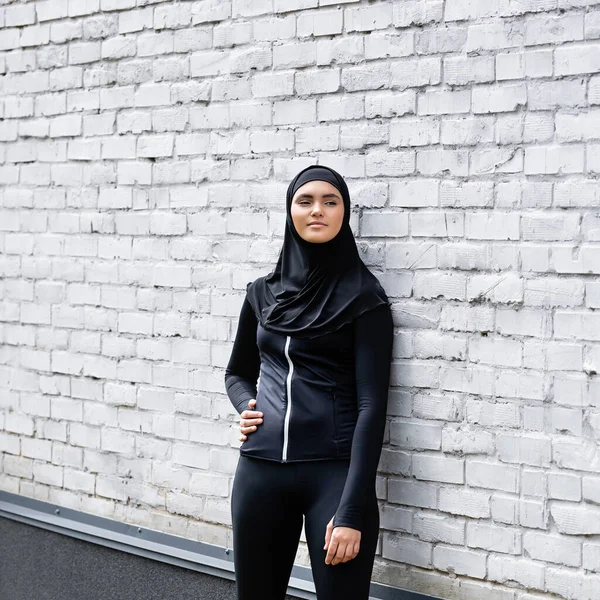 Attractive muslim girl in hijab standing with hand on hip near brick wall — Stock Photo