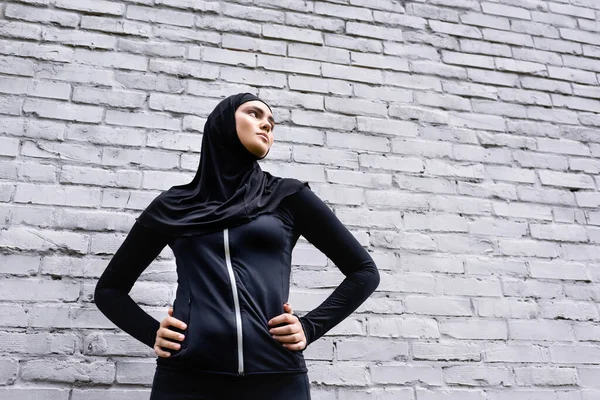 Vista de ángulo bajo de la chica musulmana en hijab de pie con las manos en las caderas cerca de la pared de ladrillo - foto de stock