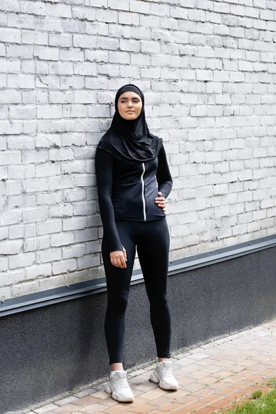 Attraente donna musulmana in hijab in piedi con mano sul fianco vicino muro di mattoni — Foto stock