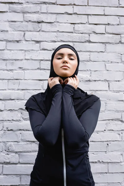 Atractiva mujer musulmana tocando hijab cerca de la pared de ladrillo - foto de stock