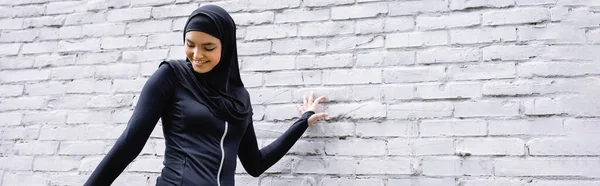Colpo panoramico di attraente donna musulmana sorridente vicino al muro di mattoni — Foto stock