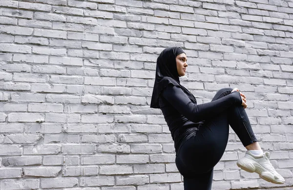 Vista de ángulo bajo de la atractiva deportista musulmana haciendo ejercicio cerca de la pared de ladrillo - foto de stock