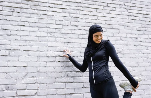 Улыбающаяся мусульманская спортсменка, тренирующаяся у кирпичной стены — стоковое фото