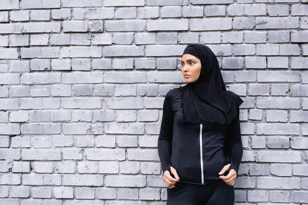 Young muslim sportswoman in hijab standing near brick wall and looking away — Stock Photo