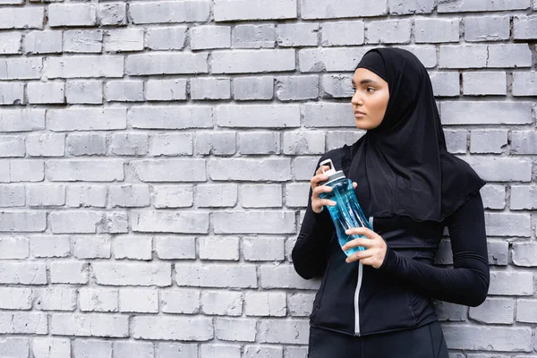 Joven deportista musulmana en hijab sosteniendo botella deportiva con agua afuera - foto de stock