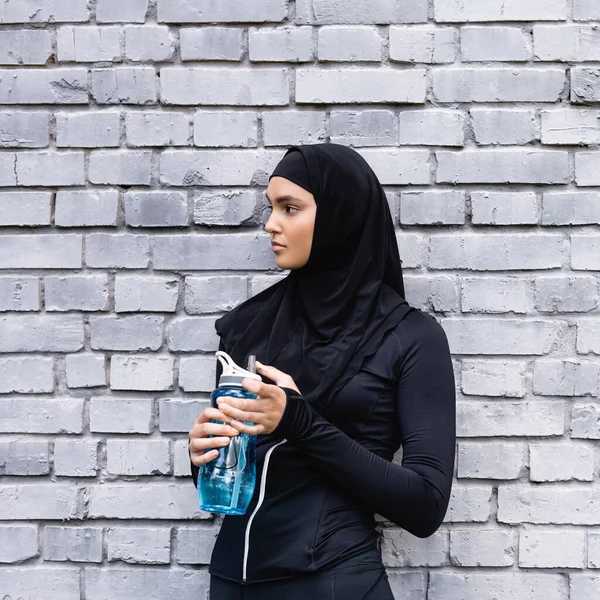Joven deportista musulmana en hijab sosteniendo botella deportiva con agua cerca de la pared de ladrillo - foto de stock
