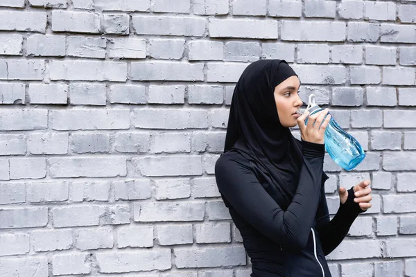 Joven deportista musulmana en hijab sosteniendo botella deportiva mientras bebe agua afuera - foto de stock