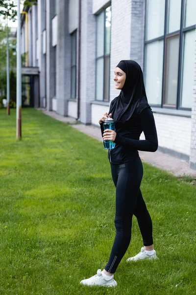 Cheerful muslim sportswoman in hijab holding sports bottle with water while standing on green grass — Stock Photo