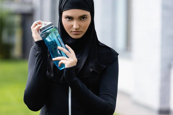 Joven deportista musulmana en hijab sosteniendo botella deportiva con agua y mirando a la cámara - foto de stock