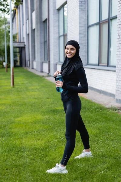 Heureuse sportive musulmane en hijab tenant bouteille de sport avec de l'eau tout en se tenant debout sur l'herbe verte — Photo de stock