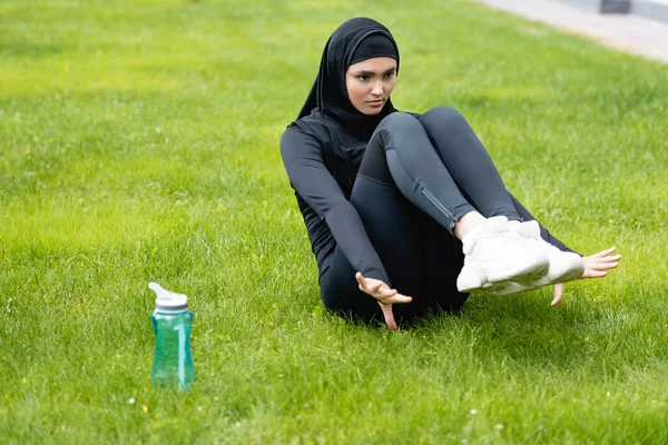 Young muslim sportswoman in hijab working out on grass near sports bottle with water — Stock Photo