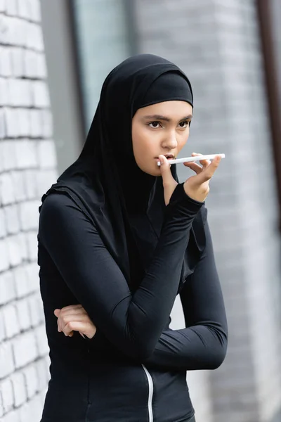 Mulher muçulmana gravando mensagem de voz e segurando smartphone perto da parede de tijolo — Fotografia de Stock