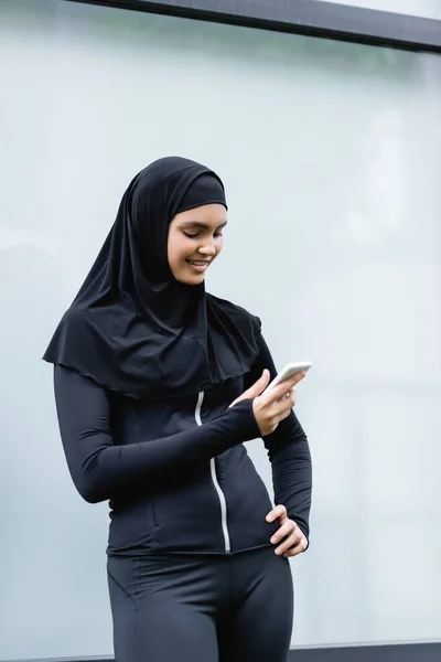 Mujer musulmana feliz usando teléfono inteligente y de pie con la mano en la cadera - foto de stock