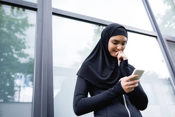 Vista de ángulo bajo de la mujer musulmana alegre mirando el teléfono inteligente - foto de stock