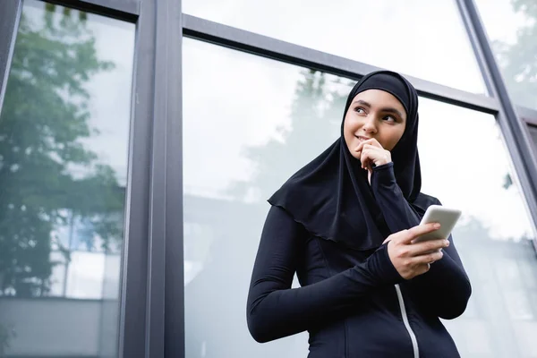 Vista de ángulo bajo de la alegre mujer musulmana sosteniendo el teléfono inteligente y mirando hacia otro lado - foto de stock