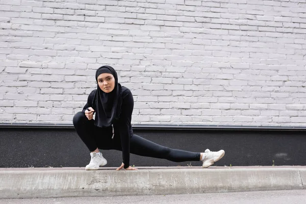 Arabian sportswoman in hijab stretching near brick wall — Stock Photo
