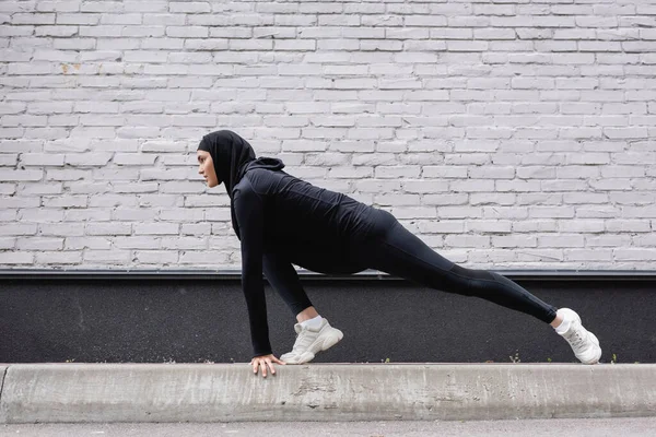 Side view of arabian sportswoman in hijab stretching near brick wall — Stock Photo