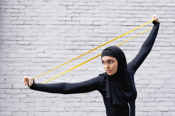 Arabian sportswoman in hijab exercising with resistance band near brick wall — Stock Photo