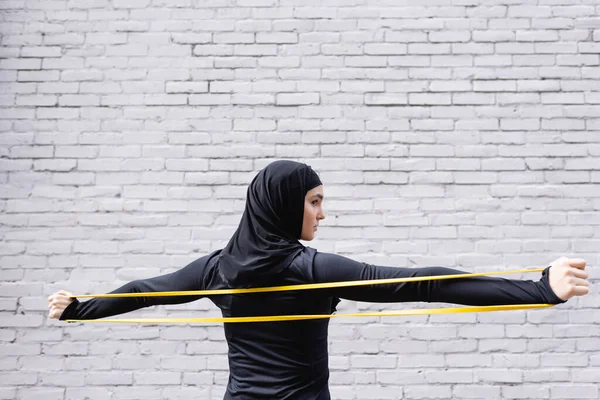 Arabian woman in hijab exercising with resistance band near brick wall — Stock Photo