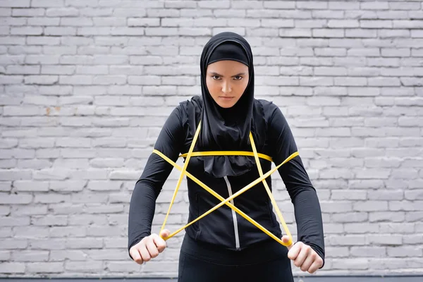 Arabian sportswoman in hijab exercising with resistance band near brick wall and looking at camera — Stock Photo