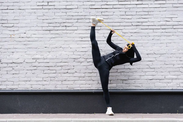 Flexible arabian sportswoman in hijab exercising with resistance band near brick wall — Stock Photo