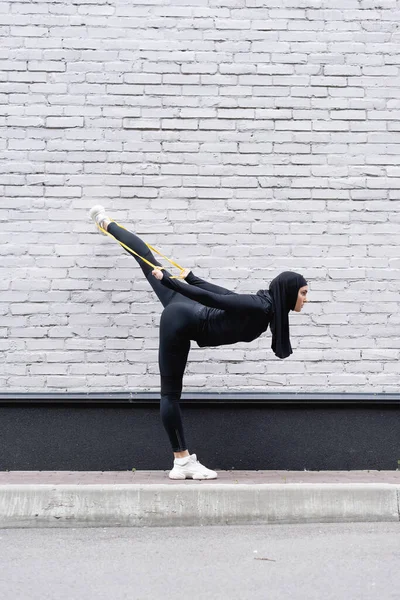 Flexible muslim sportswoman in hijab exercising with resistance band near brick wall — Stock Photo