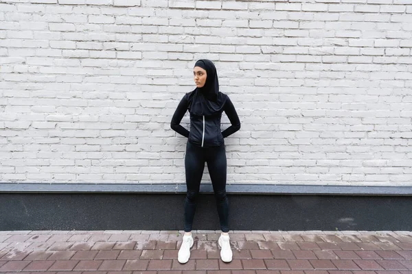 Joven deportista musulmana en hijab y ropa deportiva de pie cerca de la pared de ladrillo - foto de stock