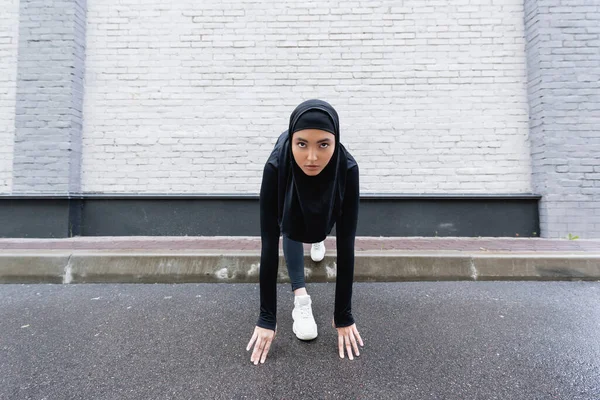 Joven deportista musulmana en hijab y ropa deportiva haciendo ejercicio cerca de la pared de ladrillo - foto de stock