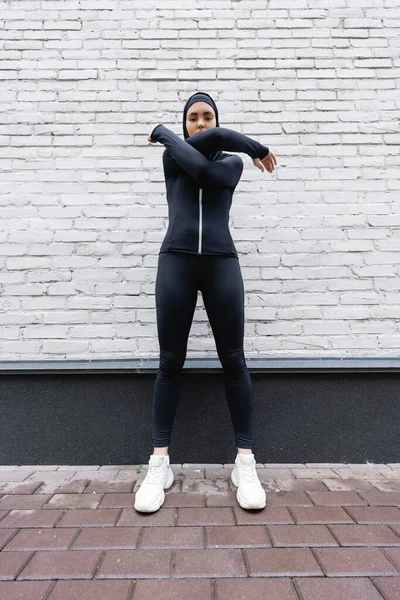 Muslim sportswoman in hijab and sportswear exercising near brick wall — Stock Photo