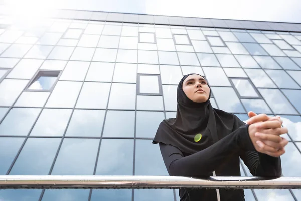 Низкий угол обзора мусульманской женщины в хиджабе, стоящей с сжатыми руками возле современного здания — стоковое фото