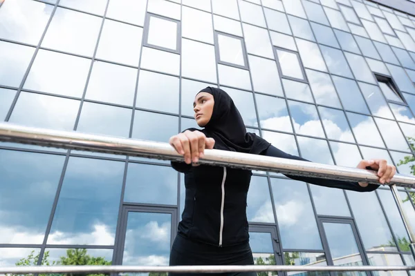 Visão de baixo ângulo da mulher árabe em hijab segurando corrimão enquanto está perto do edifício moderno — Fotografia de Stock