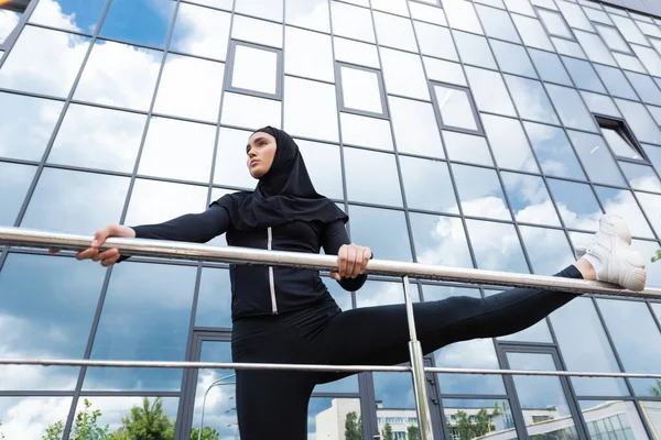 Niedrigwinkel-Ansicht der arabischen Frau im Hijab hält Geländer während der Übung in der Nähe moderner Gebäude — Stockfoto