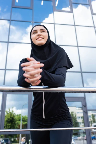 Foco seletivo da mulher muçulmana feliz em pé hijab com as mãos apertadas perto do edifício moderno — Fotografia de Stock