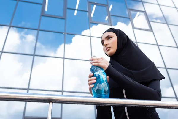 Visão de baixo ângulo da mulher muçulmana no hijab segurando garrafa de esportes perto do edifício moderno — Fotografia de Stock