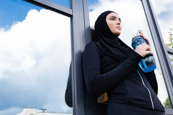 Vista de ángulo bajo de la deportista árabe en hijab celebración de botella deportiva cerca de edificio moderno - foto de stock