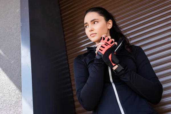 Low angle view of beautiful young woman touching sport gloves and standing outside — Stock Photo
