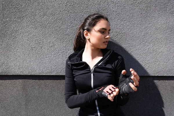 Giovane e attraente donna toccando guanti sportivi vicino muro di cemento — Foto stock