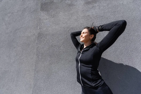 Luz del sol sobre la joven deportista feliz sonriendo cerca de la pared de hormigón - foto de stock