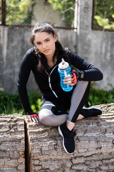 Fille en vêtements de sport tenant bouteille de sport avec de l'eau et assis sur le tronc d'arbre — Photo de stock