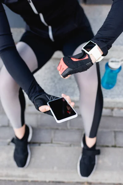 Vista recortada de la deportista con rastreador de fitness en la mano sosteniendo teléfono inteligente con pantalla en blanco - foto de stock