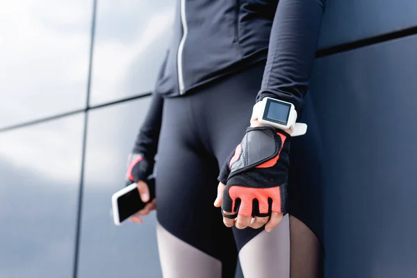 Vista recortada de la joven deportista con rastreador de fitness en la mano de pie y sosteniendo el teléfono inteligente - foto de stock