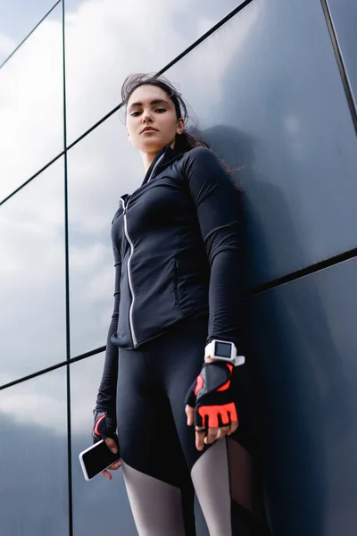 Vista de ángulo bajo de la joven deportista con rastreador de fitness en la mano de pie y la celebración de teléfono inteligente - foto de stock