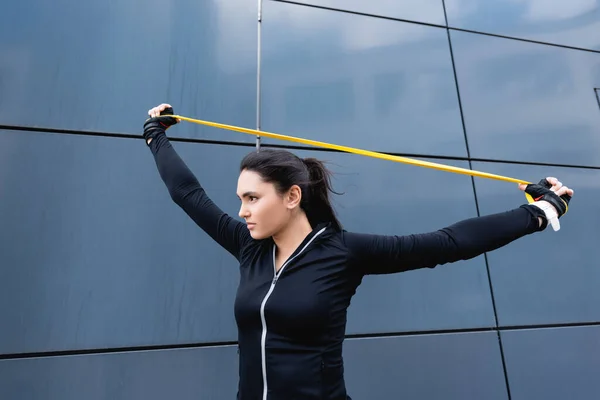 Joven deportista haciendo ejercicio con banda de resistencia cerca del edificio - foto de stock