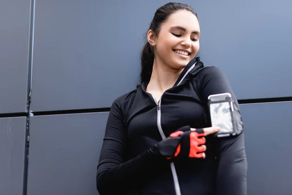 Избирательный фокус веселой спортсменки, указывающей пальцем на смартфон в браслете телефона на руке — стоковое фото