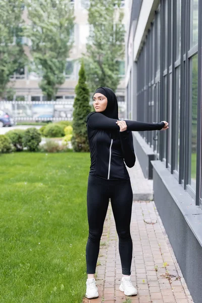 Araba donna in hijab e abbigliamento sportivo lavorando fuori vicino edificio e erba verde — Foto stock