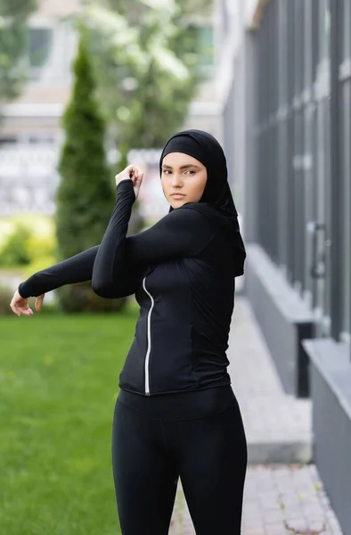 Araba ragazza in hijab e abbigliamento sportivo lavorando fuori vicino edificio e erba verde — Foto stock
