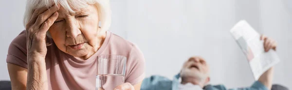 Панорамный снимок пожилой женщины, держащей стакан воды рядом с мужем с газетой, страдающей от жары дома — стоковое фото