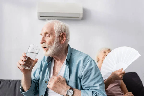 Enfoque selectivo del hombre mayor sosteniendo un vaso de agua cerca de la esposa con ventilador en el sofá - foto de stock
